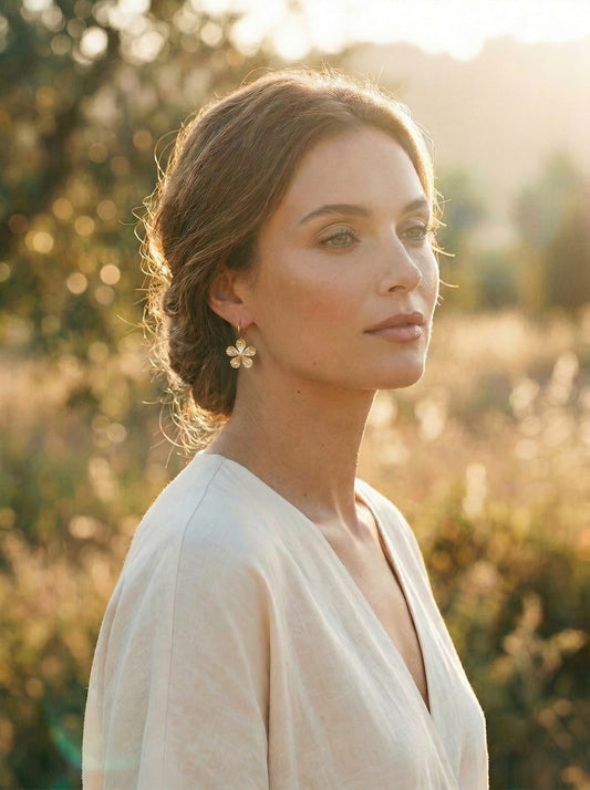 Woman in a white dress standing in a field with a warm, blurred background wearing gold flower shaped earrings