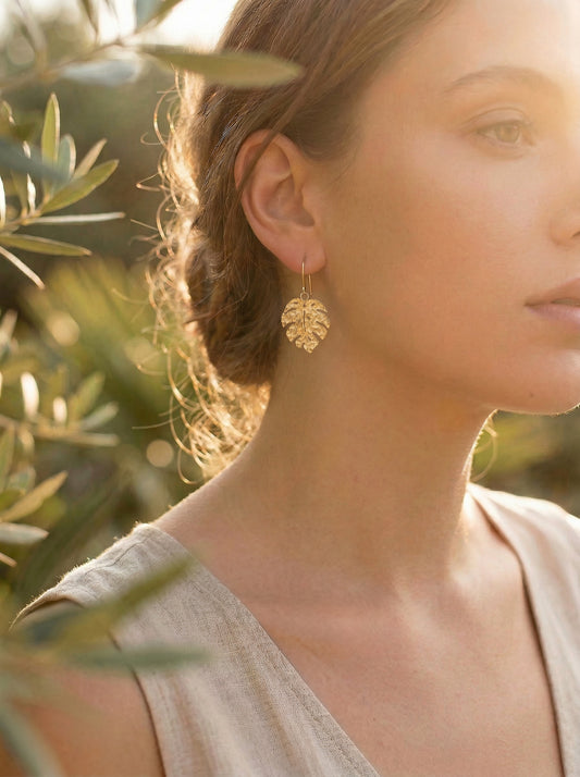 Woman wearing gold monstera leaf earrings with a blurred natural background