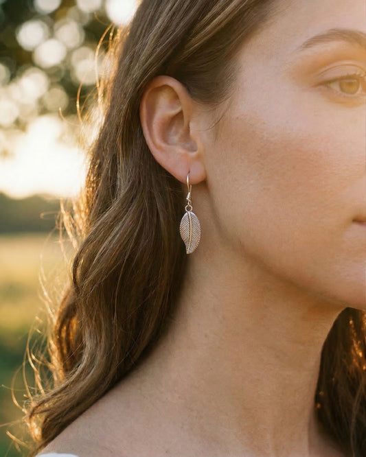 Close-up of a person wearing silver leaf-shaped earrings with a blurred natural background