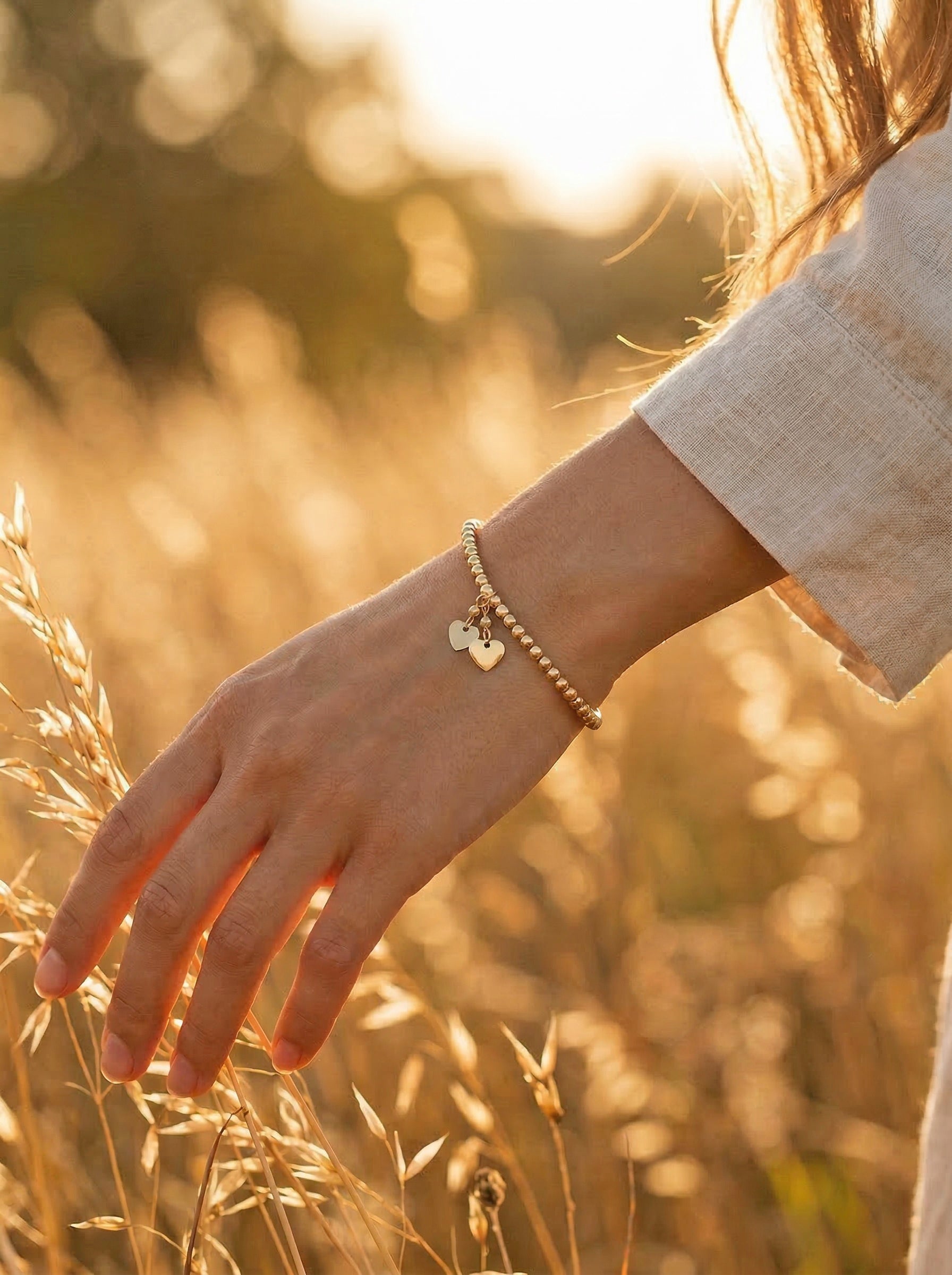 Hand wearing a gold bracelet with heart charm in a field of tall grass during sunset.