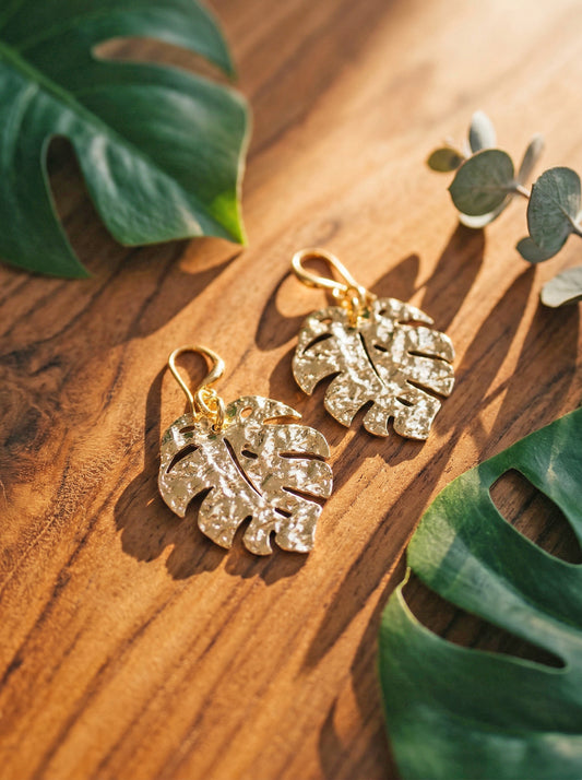 Gold monstera leaf shaped earrings on a wooden surface with green leaves.