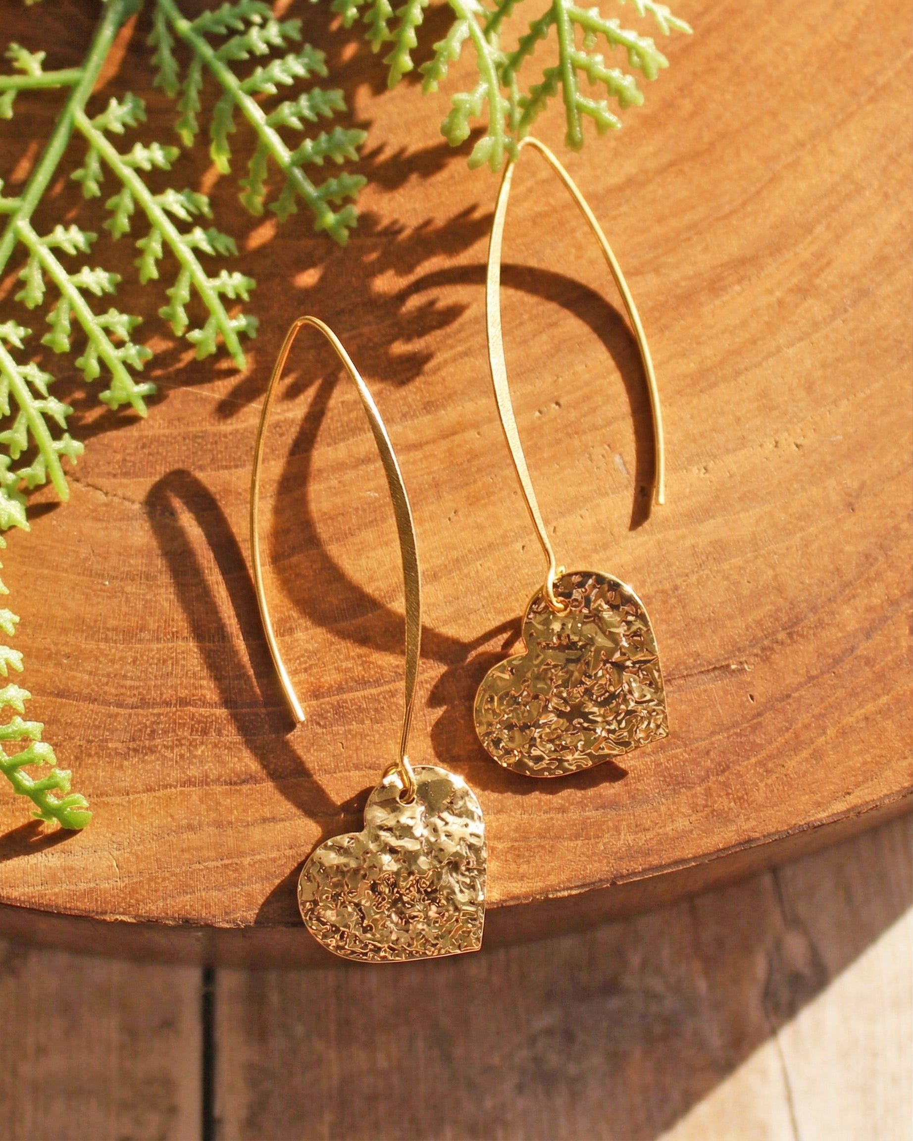 Gold heart-shaped earrings on a wooden surface with green leaves.