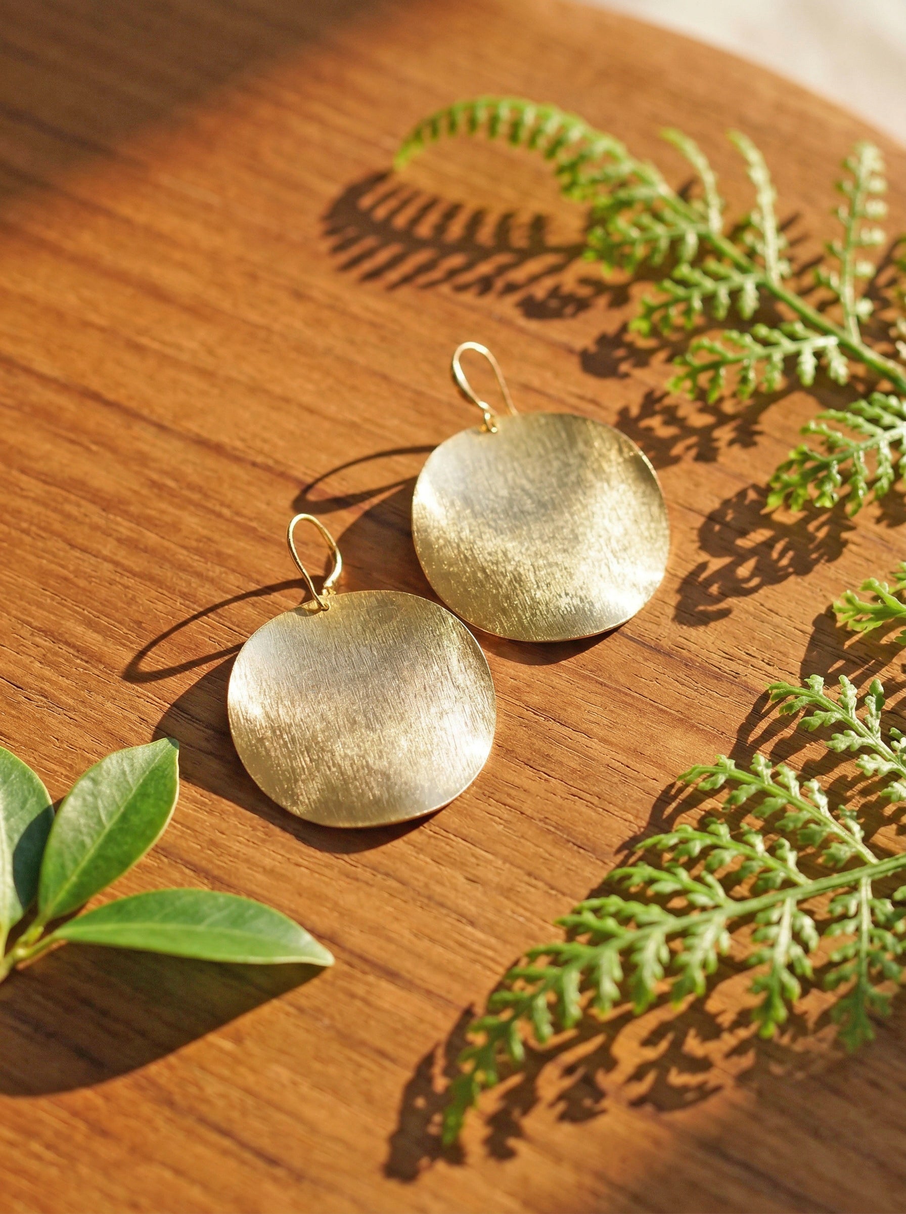 Large gold disc earrings on a wooden surface with green leaves