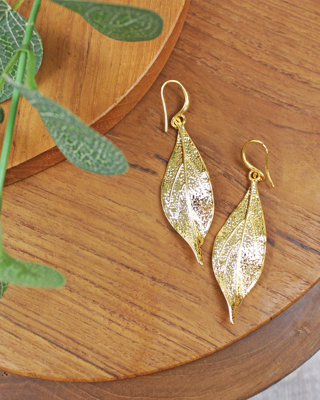 Gold leaf-shaped earrings on a wooden surface with green leaves.