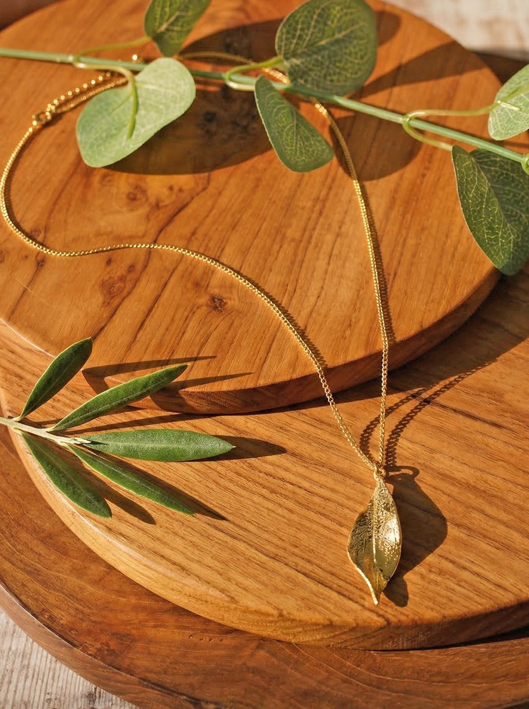 Gold necklace with a leaf pendant on a wooden surface with green leaves.