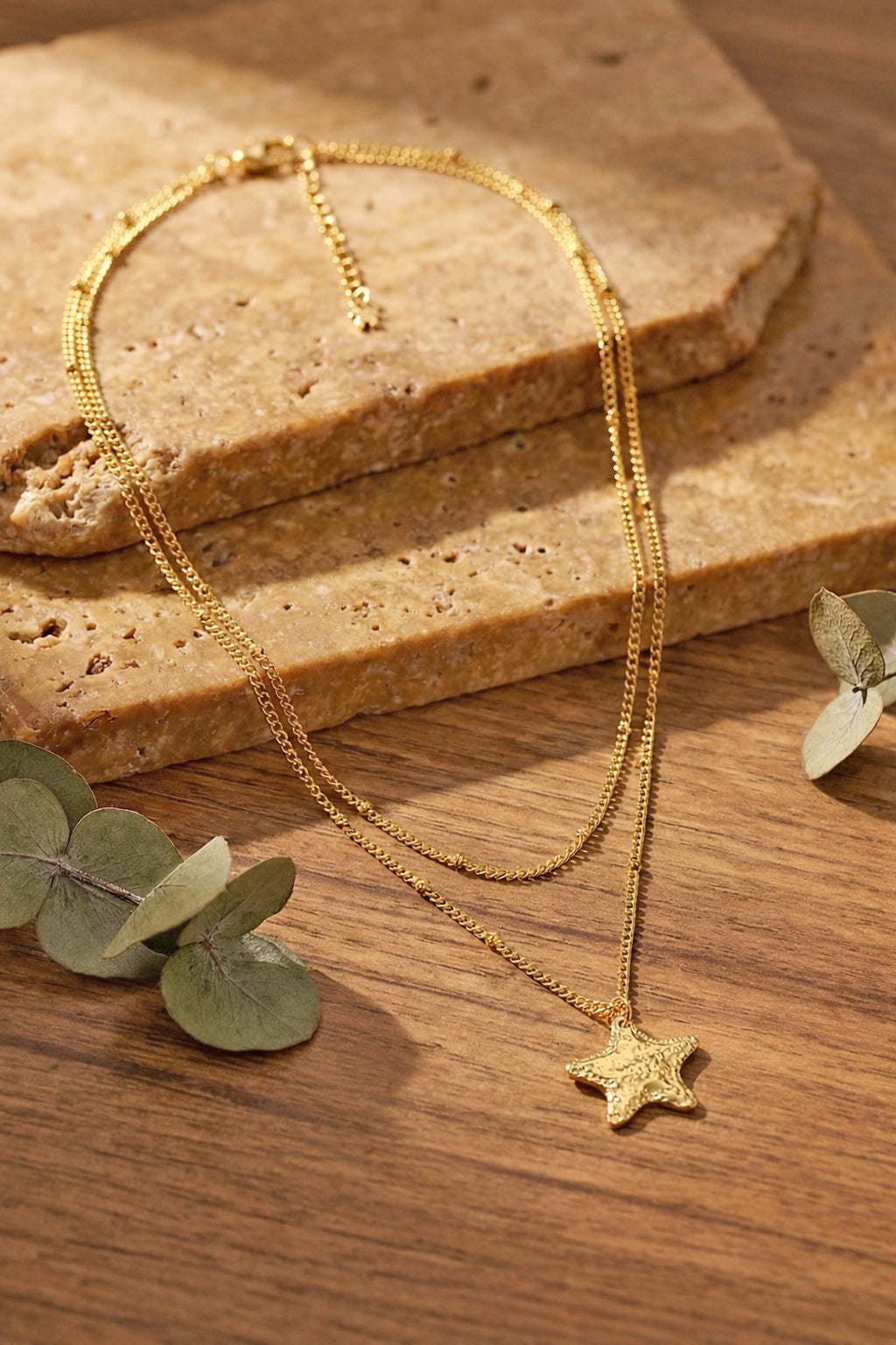 Gold-plated necklace with a star pendant on a wooden surface with eucalyptus leaves.