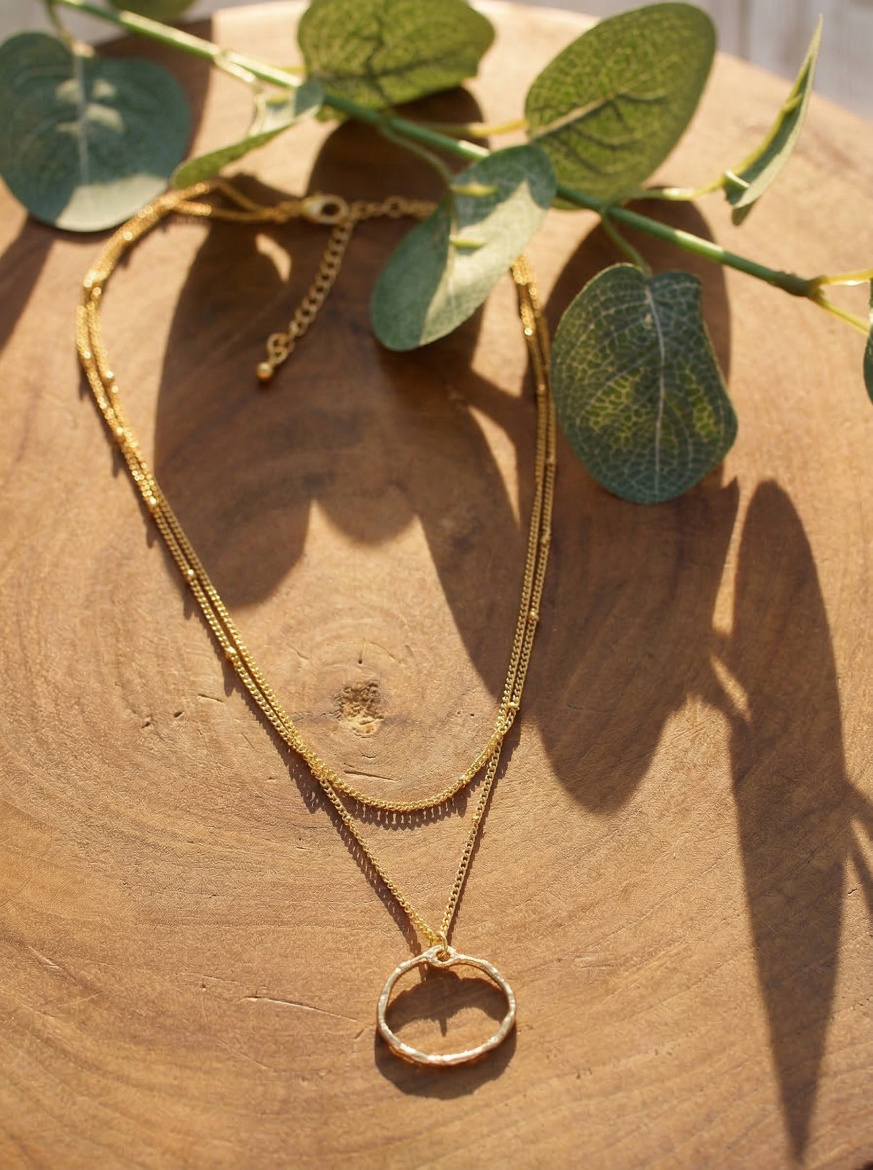 Gold-plated layered ring necklace on a wooden surface with green leaves