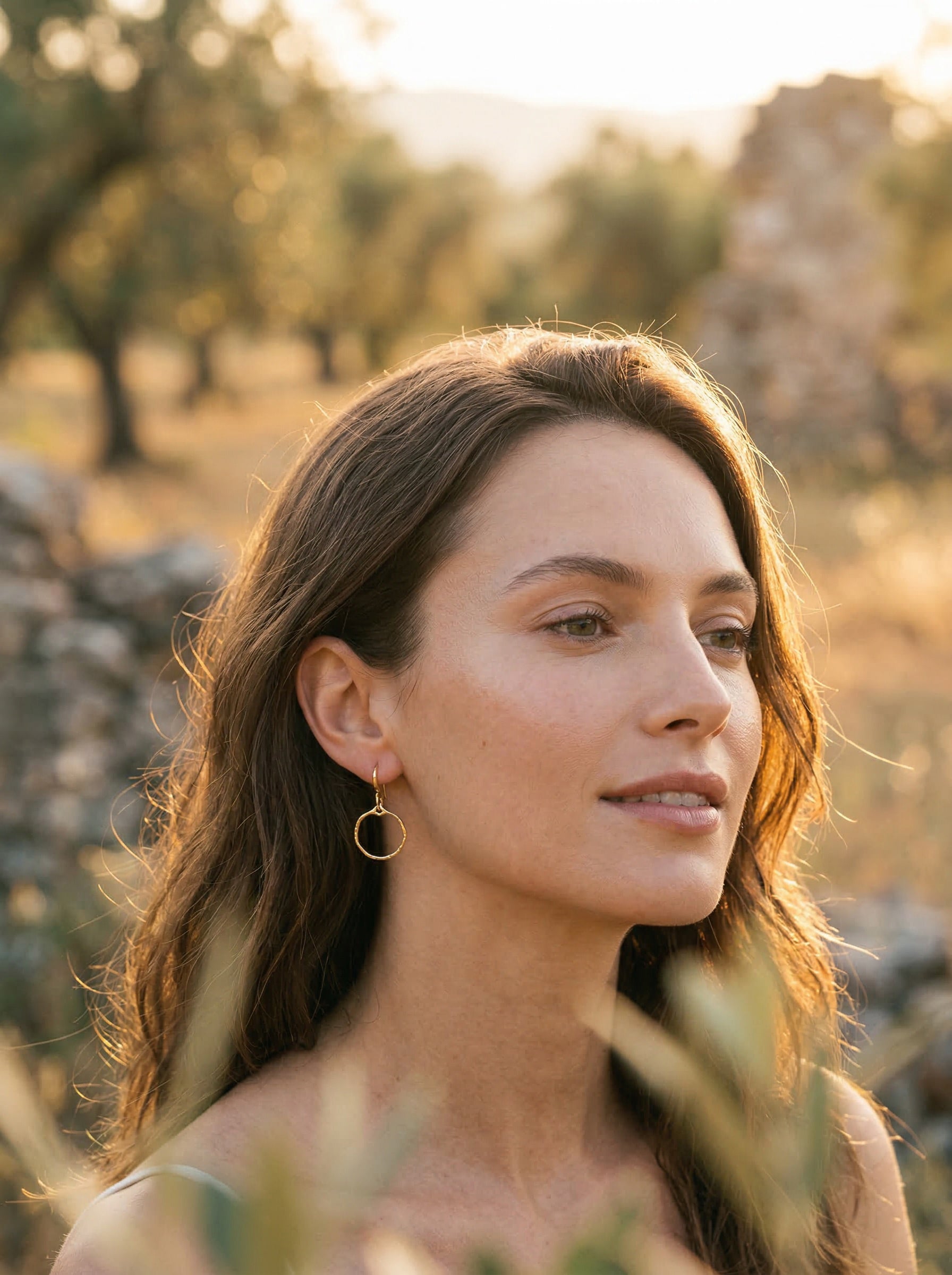 Woman with long brown hair standing in a natural setting with trees and stone structures weatring gold hoop earrings