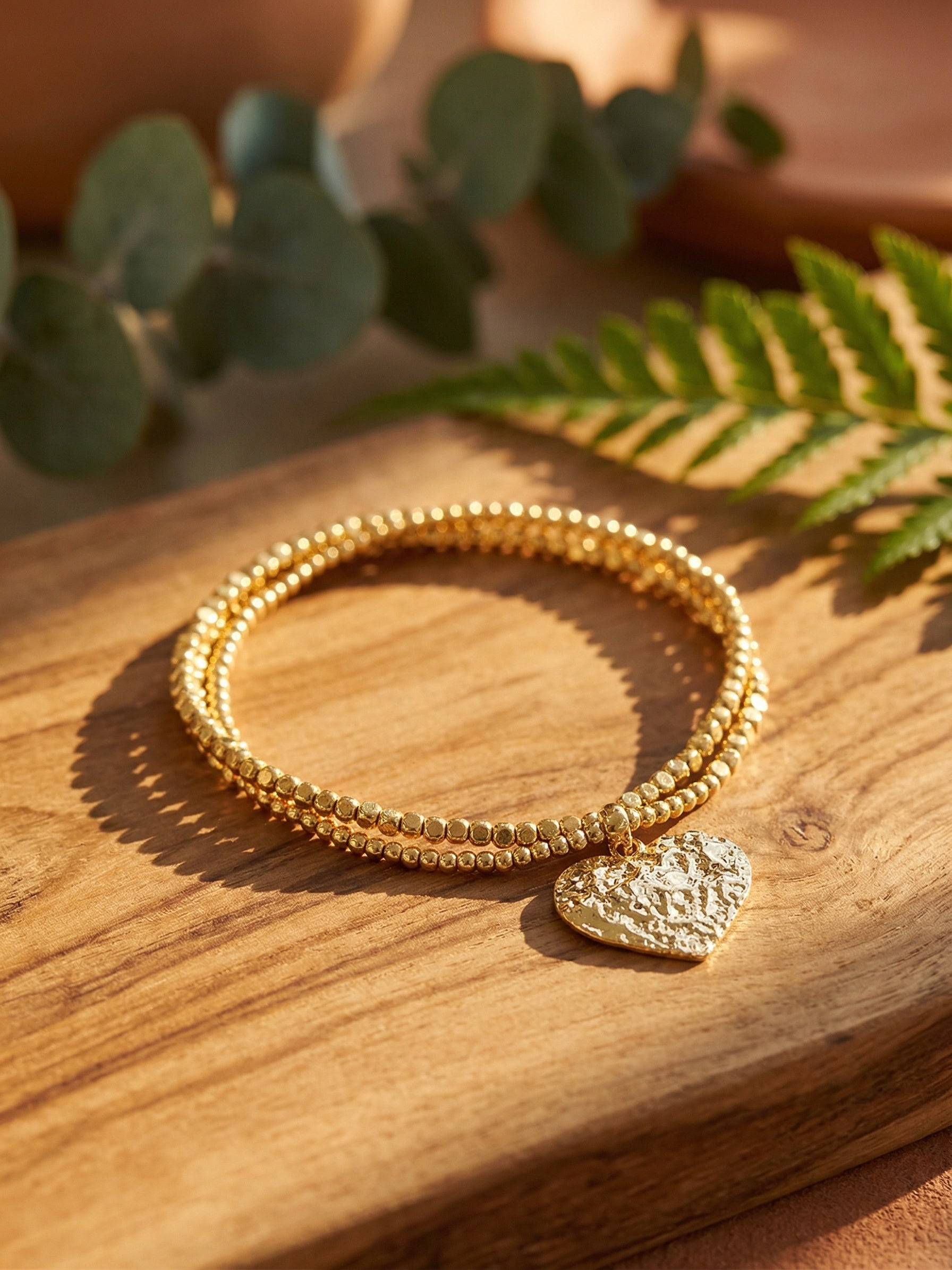 Gold bracelets with a heart charm on a wooden surface with green leaves in the background