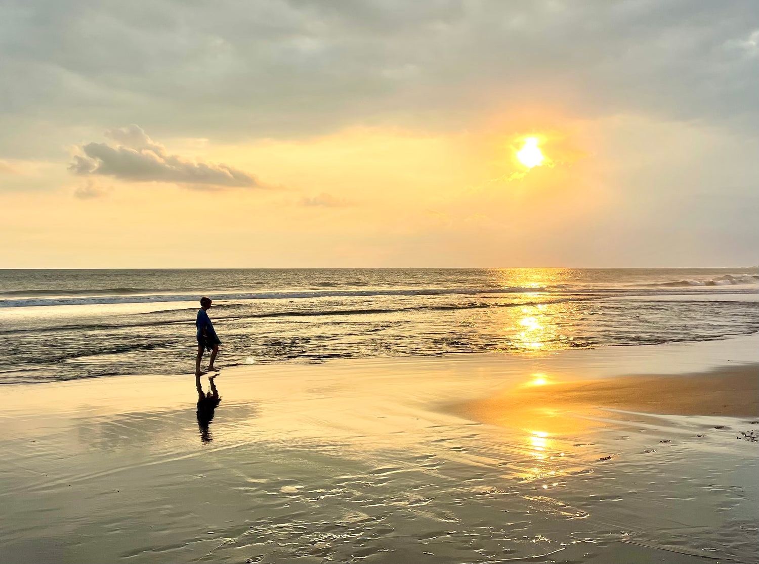 Person standing on a beach at sunset with the sun low on the horizon.