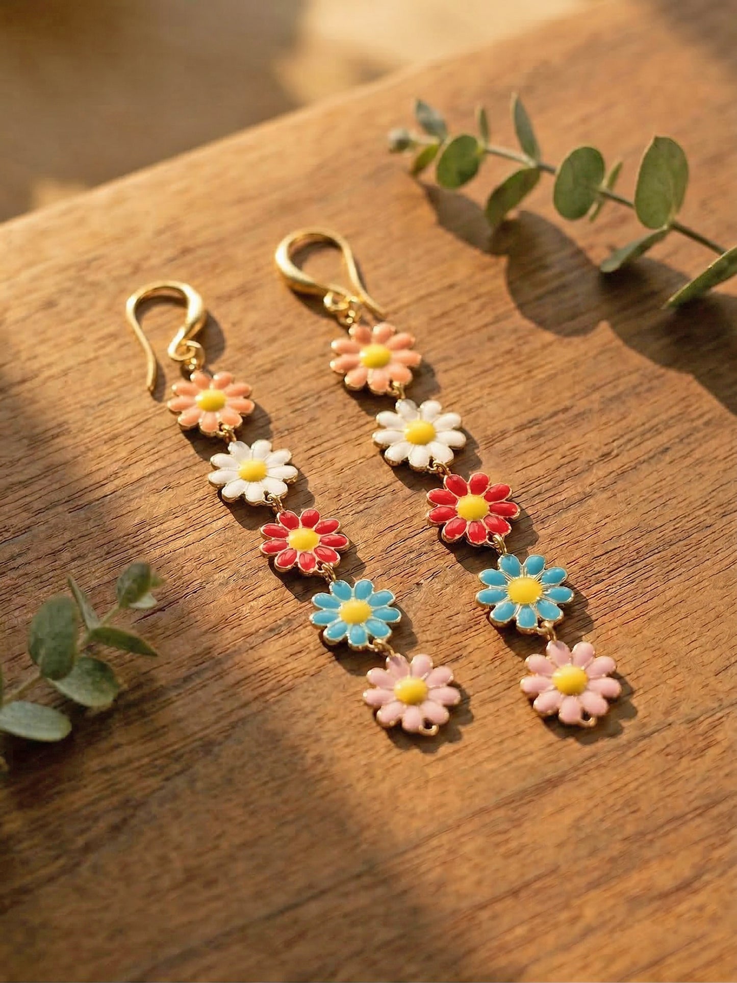 Multicoloured daisy earrings on a wooden surface with green leaves.