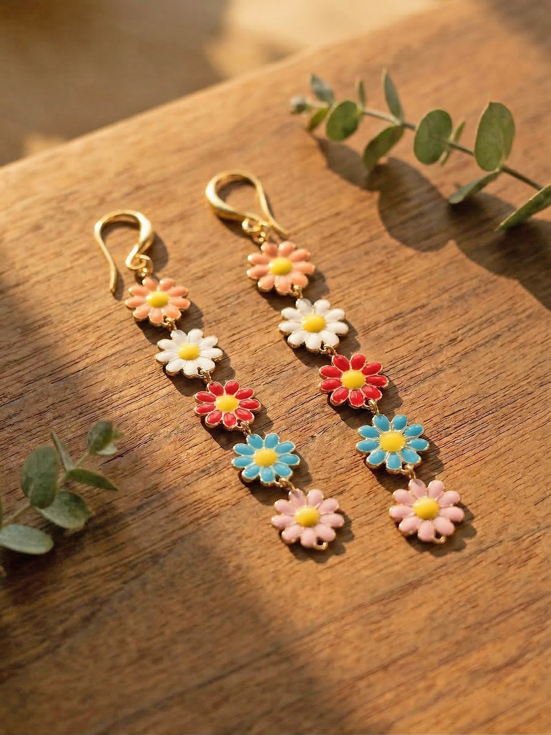 Multicoloured daisy earrings on a wooden surface with green leaves.