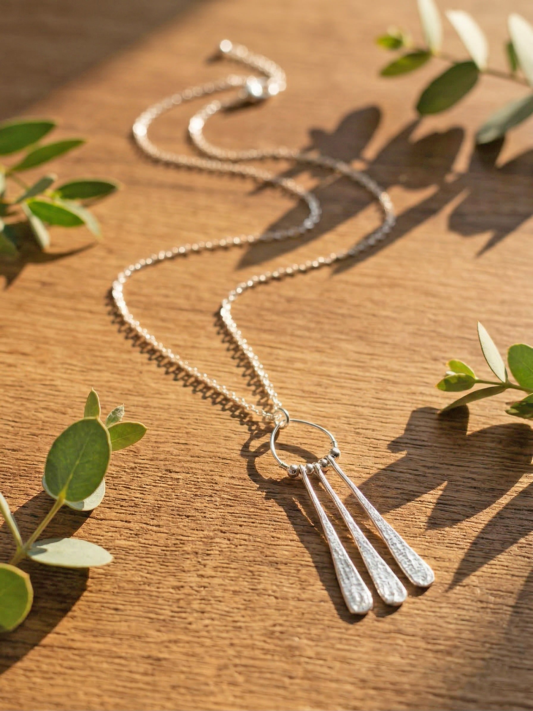 Silver necklace with three droplet pendants on a wooden surface with green leaves.