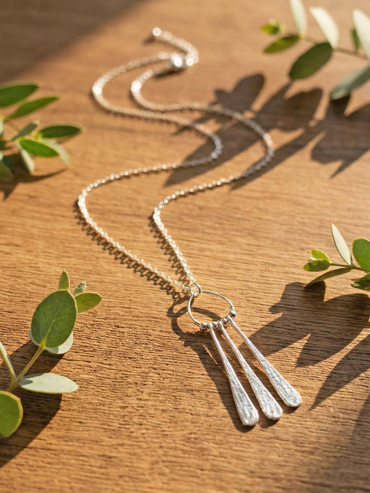 Silver necklace with three droplet pendants on a wooden surface with green leaves.