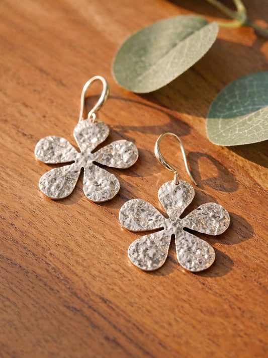 Silver flower-shaped earrings on a wooden surface with leaves in the background