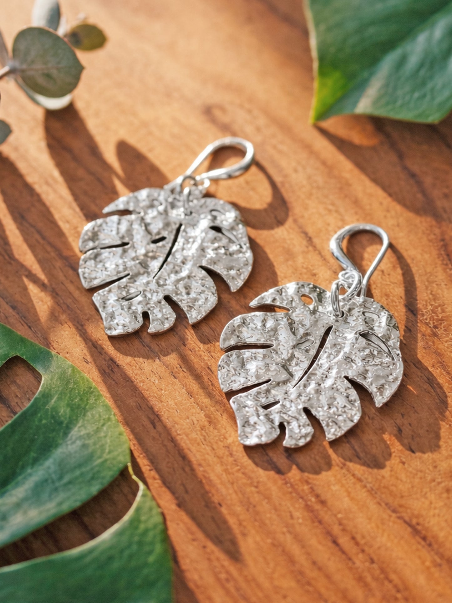 Silver monstera leaf shaped earrnigs on a wooden surface with green leaves in the background