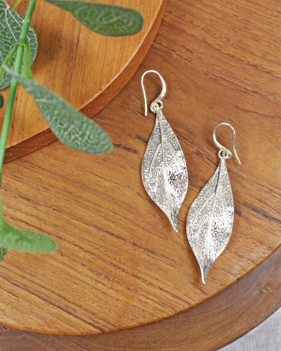 Silver leaf-shaped earrings on a wooden surface with green leaves.