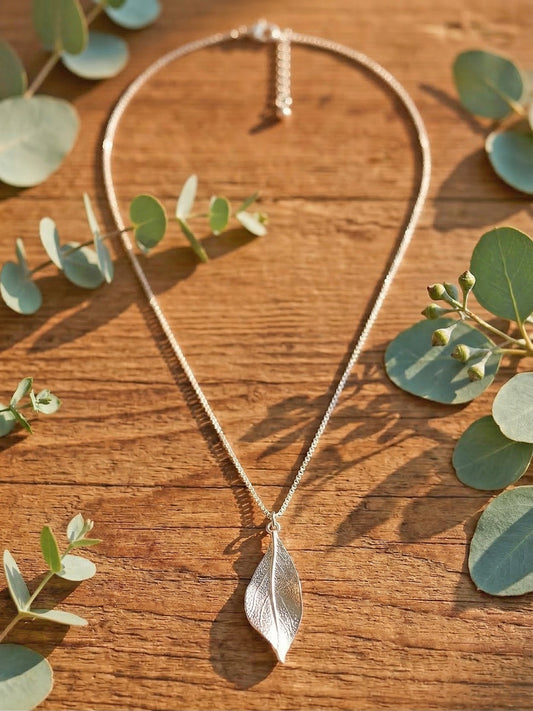 Silver leaf pendant necklace on a wooden surface with eucalyptus leaves