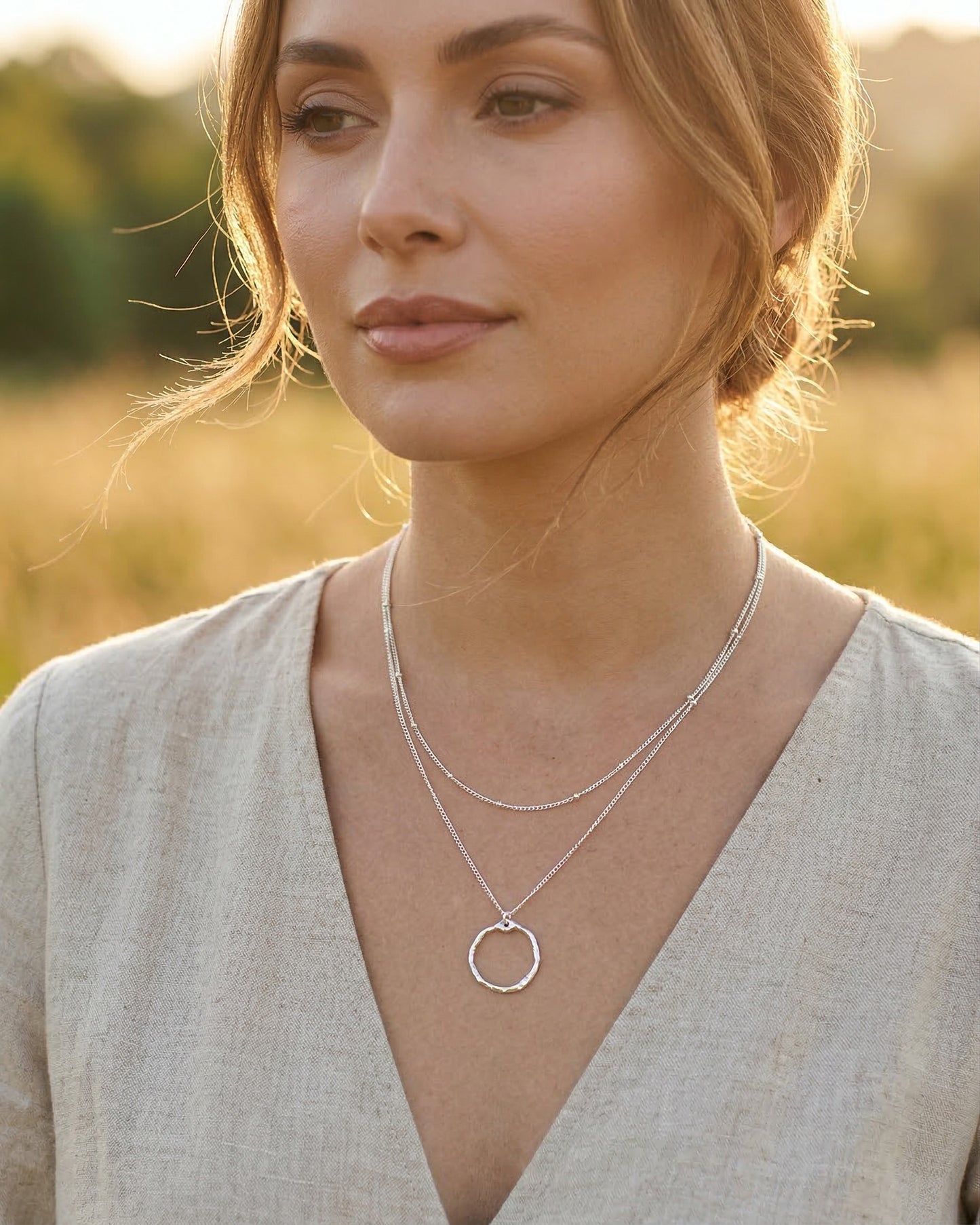 Woman wearing a silver layered necklace with a circular pendant in a field at golden hour
