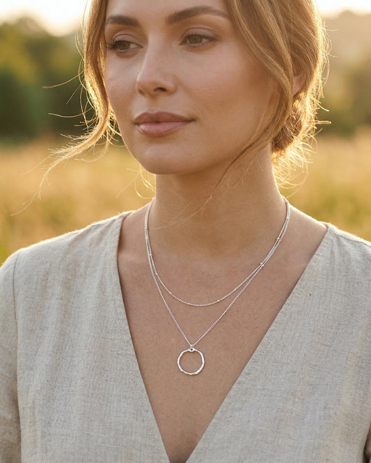 Woman wearing a silver layered necklace with a circular pendant in a field at golden hour