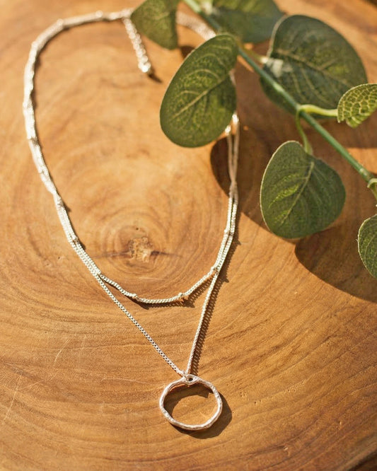 Silver layered necklace with a circular pendant on a wooden surface with green leaves.