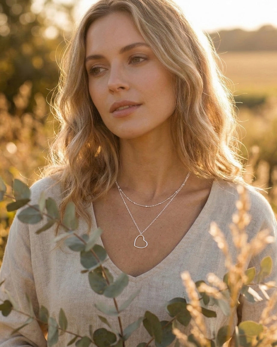 Woman wearing a heart-shaped layered necklace in a natural setting with greenery and golden sunlight