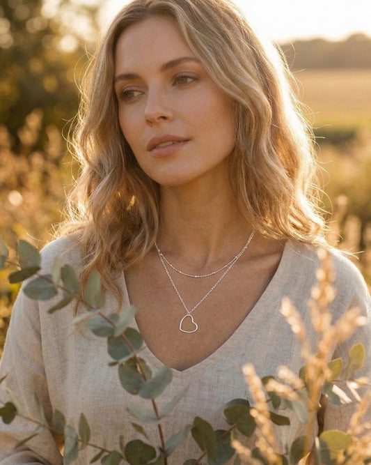 Woman wearing a heart-shaped layered necklace in a natural setting with greenery and golden sunlight