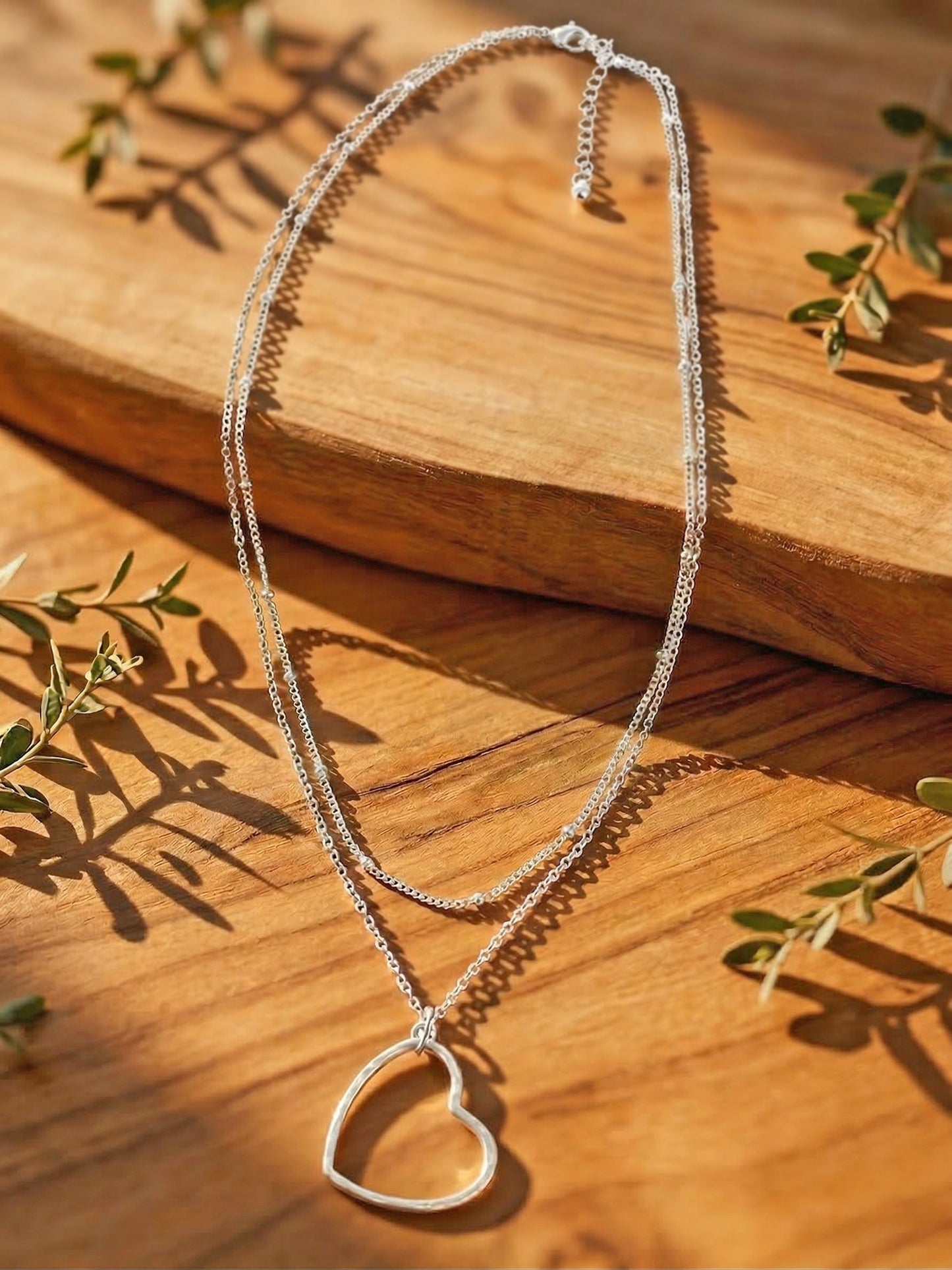 Silver heart-shaped layered necklace on a wooden surface with green leaves