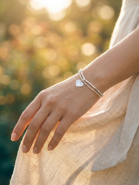 Hand wearing a silver bracelet with a heart charm against a blurred natural background