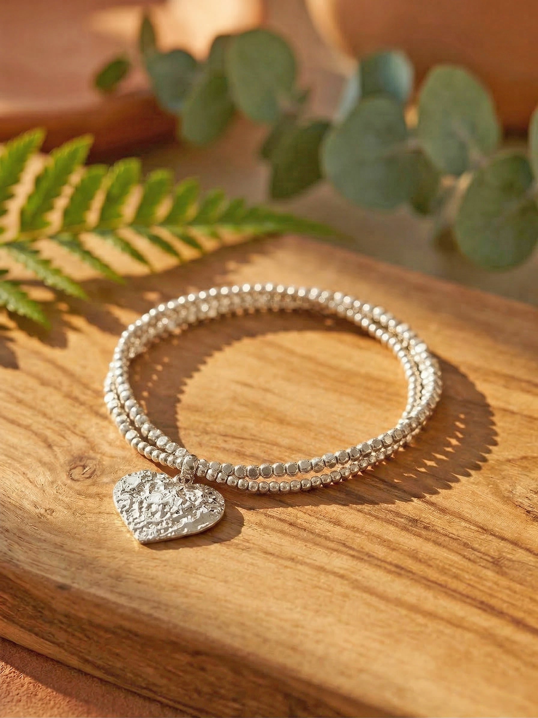 Silver bracelet with a heart charm on a wooden surface with green leaves in the background