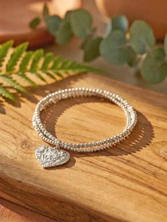 Silver bracelet with a heart charm on a wooden surface with green leaves in the background