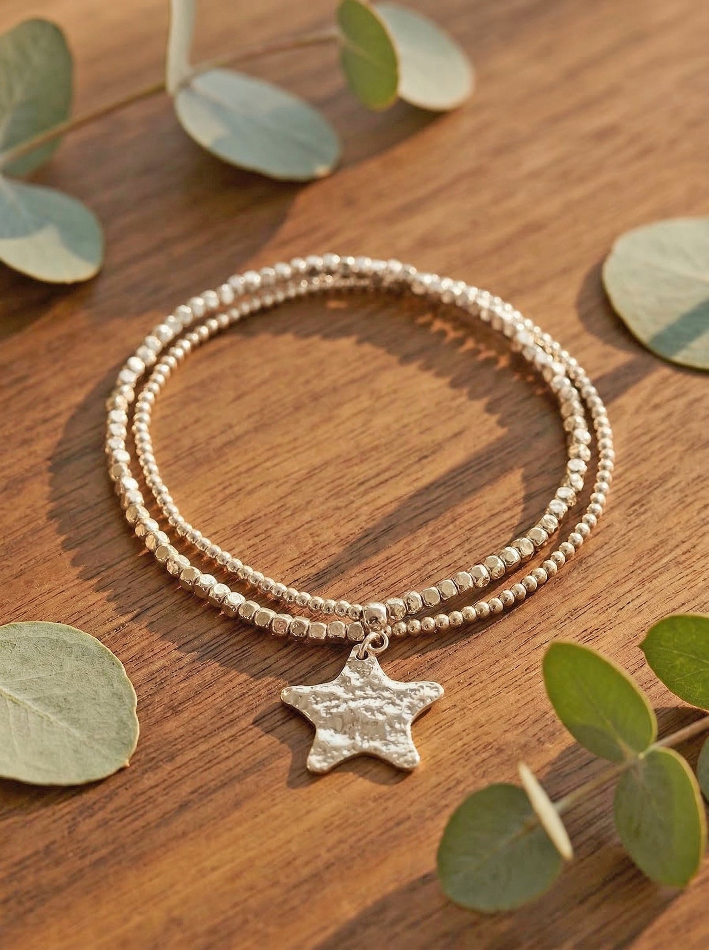 Silver beaded bracelet with a star charm on a wooden surface with eucalyptus leaves.
