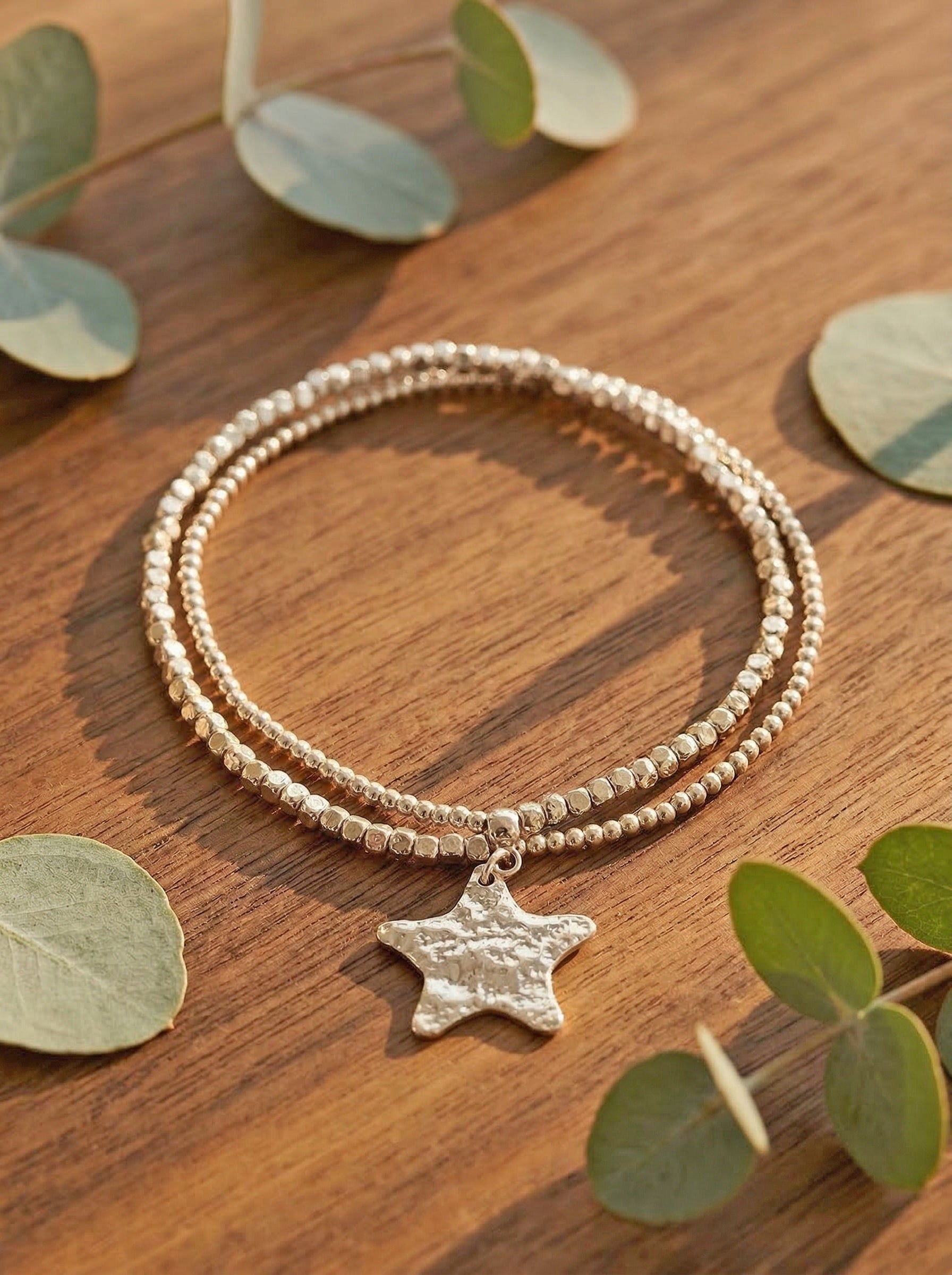 Silver beaded bracelet with a star charm on a wooden surface with eucalyptus leaves.