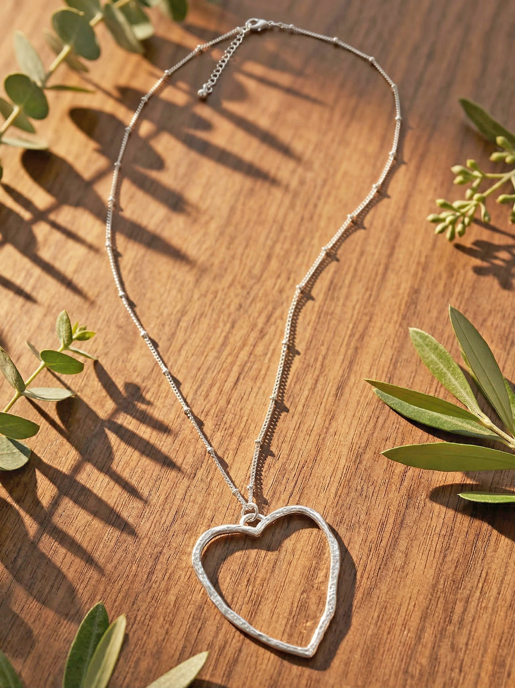 Silver heart-shaped necklace on a wooden surface with green leaves