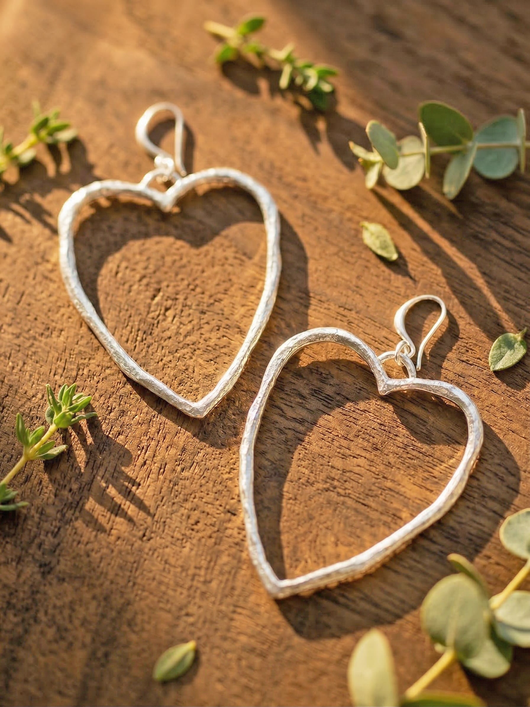 Heart-shaped silver earrings on a wooden surface with green leaves.