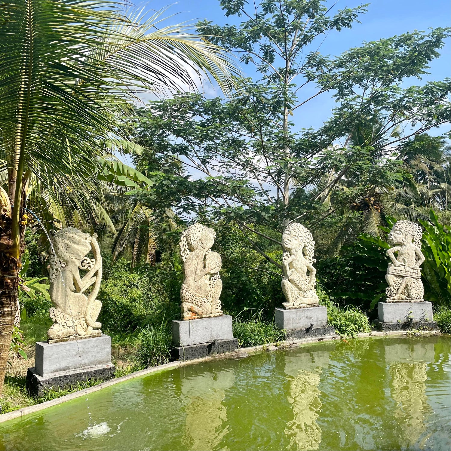 Four stone statues by a pond with greenery in the background